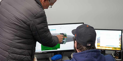 Two men are staring at a computer screen with a 3D model of a pump on it. The man on the left is pointing to part of the diagram/illustration on the computer.